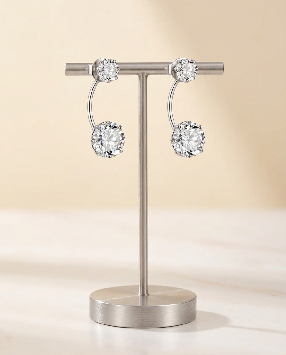 Double drop earrings in polished sterling silver with a small round clear stone above a larger one, on a silver T-bar stand against a warm ivory backdrop
